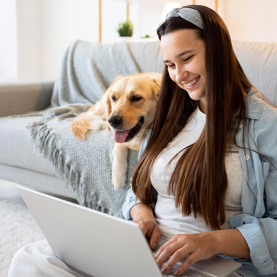 woman-on-laptop-with-dog-nearby A smiling young woman sits by her dog and looks at her computer