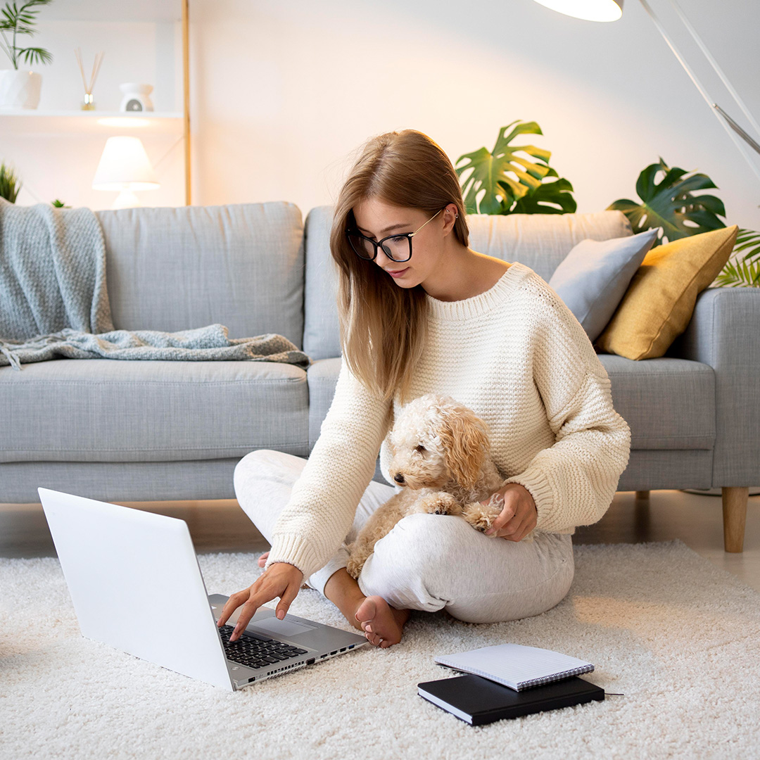 A woman sits cross legged on the floor with a small dog in her lap while she works on her computer